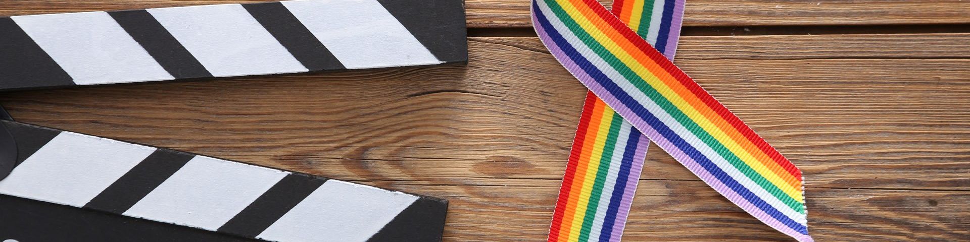 On the left is the black and white striped top of a clapperboard. Beside it is a Pride rainbow ribbon. They are both sat on a wooden table.